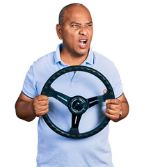 Hispanic middle age man holding steering wheel angry and mad screaming frustrated and furious,...