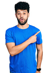 Young arab man with beard wearing casual blue t shirt surprised pointing with finger to the side, open mouth amazed expression.