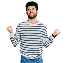 Fototapeta premium Young arab man with beard wearing casual striped sweater very happy and excited doing winner gesture with arms raised, smiling and screaming for success. celebration concept.