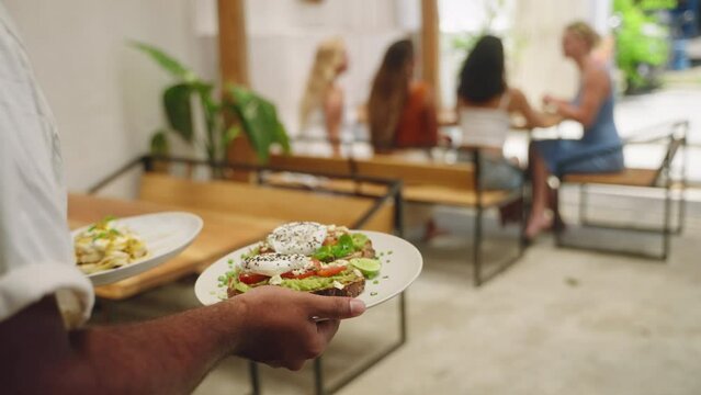 Waiter hands carry food, avocado toasts with poached egg on plates for female customers in cafe. Waiter serving food to clients. Diverse young women eat tasty dishes in the restaurant. Close-up.