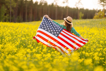 Beautiful young woman holding an American flag in the wind in a field. Summer landscape against the...