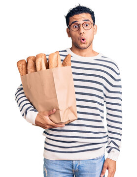 Hispanic handsome young man holding paper bag with bread scared and amazed with open mouth for surprise, disbelief face