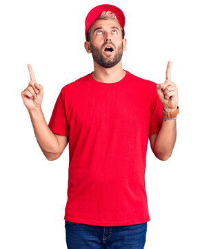 Young handsome blond man wearing t-shirt and cap amazed and surprised looking up and pointing with fingers and raised arms.