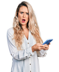 Young blonde woman using smartphone in shock face, looking skeptical and sarcastic, surprised with open mouth