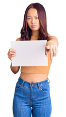 Fototapeta premium Young beautiful chinese girl holding paper banner with blank space pointing with finger to the camera and to you, confident gesture looking serious