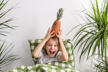 The young boy, about 9-10 years old, lies in bed, his long hair flowing, cradling a pineapple,...