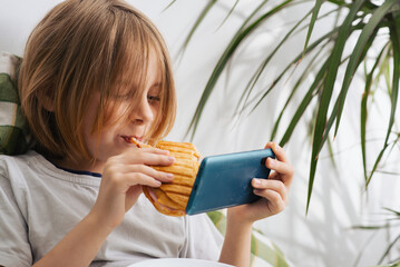 In the bed, a 9-10-year-old boy with lengthy hair savors a toast, leisurely using his mobile phone,...