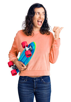 Young beautiful hispanic woman holding skate pointing thumb up to the side smiling happy with open mouth