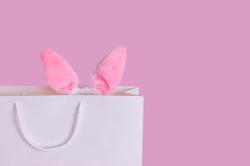 Close up of pink Easter bunny ears in a white paper bag on a pink background.