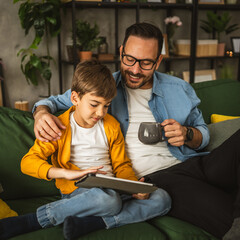 Father hold cup of coffee and look son while he use tablet for fun