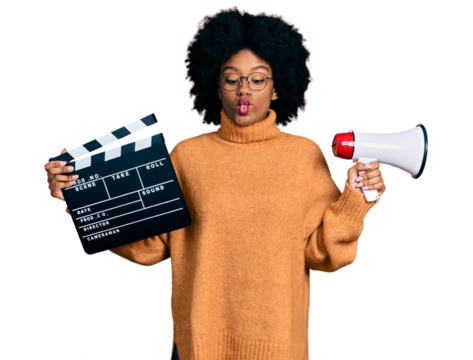 Young african american woman holding video film clapboard and megaphone making fish face with mouth and squinting eyes, crazy and comical.