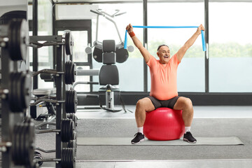 Mature man in sportswear sitting on a fitness ball and exercising with an elastic band at a gym