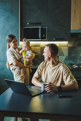 A young couple with a child in the kitchen. A man works at home on a laptop, and his wife with a...