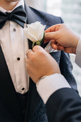 Groom with a white rose boutonniere in a classic suit close-up. The groom's friend helps the groom to put on the boutonniere. The groom and best man are preparing for the wedding