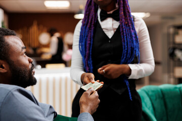 Young African American man hotel guest giving cash to waitress while relaxing in lobby, ordering...