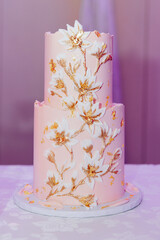 A beautiful delicate two-tiered holiday cake is decorated with white and gold confectionery flowers. Peach-colored wedding cake close-up. Concept of elegant holiday desserts