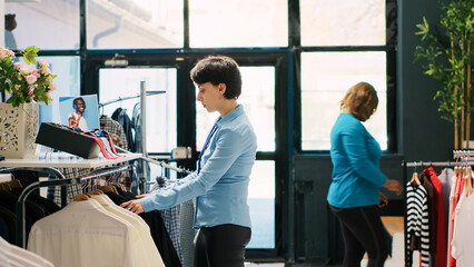 Cheerful manager working at boutique visual, putting hangers with new fashion collection on racks....