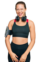 Beautiful blonde woman wearing gym clothes and using headphones winking looking at the camera with...