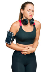 Beautiful blonde woman wearing gym clothes and using headphones with hand on stomach because indigestion, painful illness feeling unwell. ache concept.