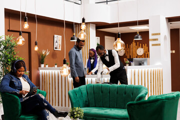 Friendly smiling African American guy bellhop helping guest with luggage during arrival at resort hotel. Happy black woman tourist in headphones using digital tablet while relaxing at reception area