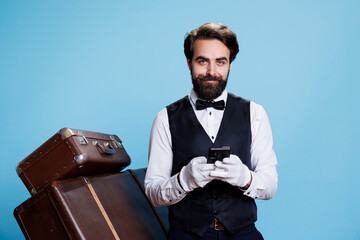 Hotel porter scrolls on smartphone, using social media and texting messages while he poses in...