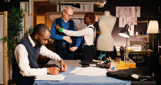 Old customer dressing up suit jacket in tailoring workshop, fitting clothes with custom made body measurements. Male client collaborating with couturiers team in fashion atelier. - Powered by Adobe