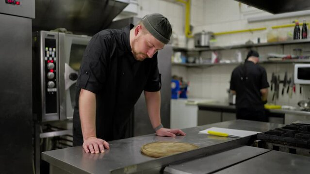 Tired male professional chef in a black uniform takes off his work cap and tries to relax during a hard day of work in the kitchen of a restaurant