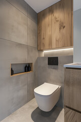 Modern bathroom with wall-mounted toilet, gray tile walls and wood cabinets above the sink. There is an integrated flush button on one of the cabinet doors. The floor has light-colored concrete tiles.
