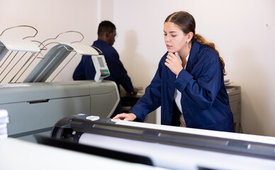 Concentrated young female typographer in a blue robe uniform pressing the buttons on the plotter in...