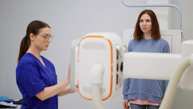 A confident brunette girl in round glasses. A doctor in a blue uniform does fluorography for a brunette girl in a jacket in a modern clinic