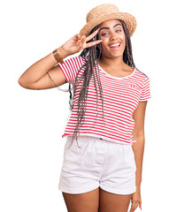 Young african american woman with braids wearing summer hat doing peace symbol with fingers over face, smiling cheerful showing victory