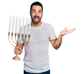 Young hispanic man with beard holding menorah hanukkah jewish candle celebrating achievement with happy smile and winner expression with raised hand