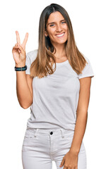 Obraz premium Young woman wearing casual white t shirt smiling with happy face winking at the camera doing victory sign. number two.