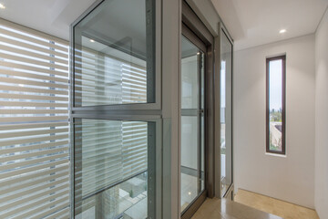 The corridor of a Mediterranean villa with light walls, a window and a glass elevator.