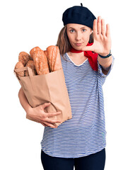Young beautiful blonde woman wearing french beret holding bag with bread with open hand doing stop...