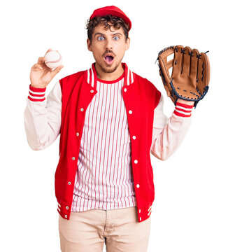 Young handsome man with curly hair wearing baseball uniform holding golve and ball scared and amazed with open mouth for surprise, disbelief face