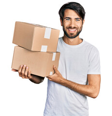 Hispanic man with blue eyes holding packages looking positive and happy standing and smiling with a...
