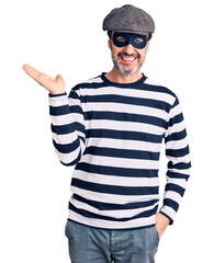 Middle age handsome man wearing burglar mask smiling cheerful presenting and pointing with palm of hand looking at the camera.