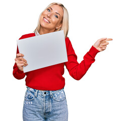 Young blonde woman holding blank empty banner smiling happy pointing with hand and finger to the side