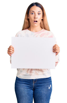 Beautiful young woman holding blank empty banner scared and amazed with open mouth for surprise, disbelief face