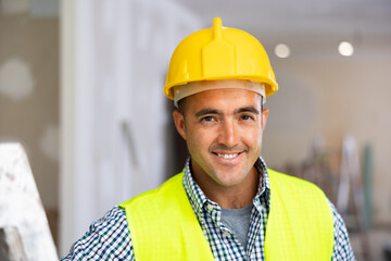 Portrait of positive man builder in vest and helmet standing in apartment during repair works,...