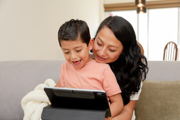 Hispanic mom and her son sitting on the couch at home using a digital tablet while having fun - mom and son watching or playing on the digital tablet - family quality time - single mother