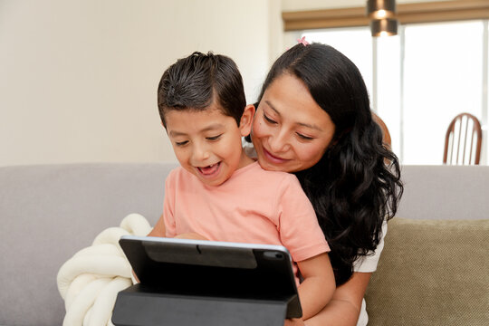Hispanic mom and her son sitting on the couch at home using a digital tablet while having fun - mom and son watching or playing on the digital tablet - family quality time - single mother