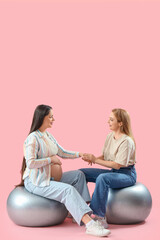 Mature doula with pregnant woman on fitballs holding hands against pink background