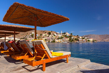 Luxurious sun loungers under reed umbrellas overlooking the scenic harbor of Symi Island, Greece