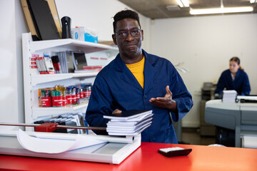 Excited middle-aged African American male typographer in uniform showing notepads in the typography