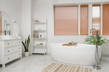 Interior of light bathroom with bathtub, chest of drawers and houseplants