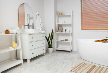 Interior of light bathroom with bathtub, chest of drawers and houseplant