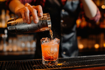Male bartender pouring a cocktail from shaker into a glass in a modern bar with blurred bar background - Powered by Adobe