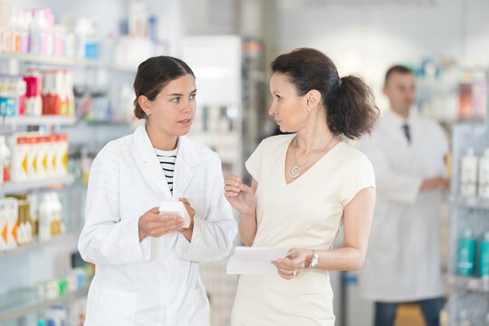 Polite young female pharmacist consulting woman client about medicament in box according to her recipe in chemist's shop